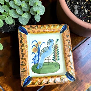 May include: A rectangular ceramic tray featuring a hand-painted light blue bird standing on a green oval patch. The center is off-white, surrounded by a textured border in shades of brown and tan with dark blue accents at each corner.