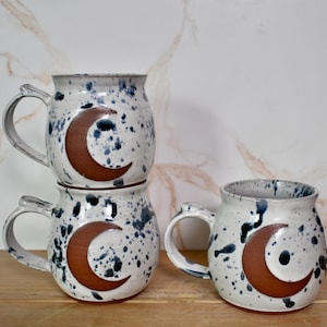 Puede incluir: Tazas de cerámica con esmalte blanco moteado y un diseño de media luna marrón. Las tazas tienen un asa resistente y están apiladas y dispuestas sobre una superficie de madera. Altura aproximada de 10 cm. Adecuadas para café, té u otras bebidas.