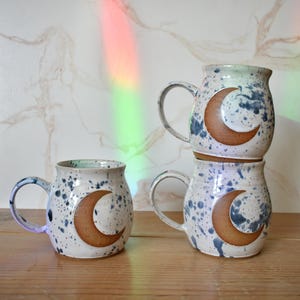 Könnte beinhalten: Vier Keramikbecher mit einer gesprenkelten weißen Glasur und einem braunen Halbmond-Design. Die Becher haben große Henkel und sind mit blauen Spritzern verziert. Die Becher sind gestapelt und auf einer Holzoberfläche angeordnet.