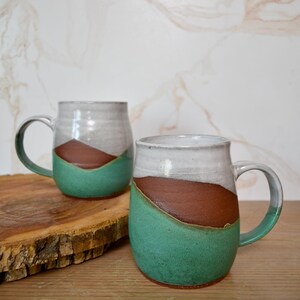 Puede incluir: Dos tazas de cer&aacute;mica hechas a mano con un dise&ntilde;o &uacute;nico. Cada taza presenta una parte superior blanca, una secci&oacute;n media marr&oacute;n y una base verde turquesa. Las tazas tienen asas grandes y se exhiben sobre una superficie de madera.