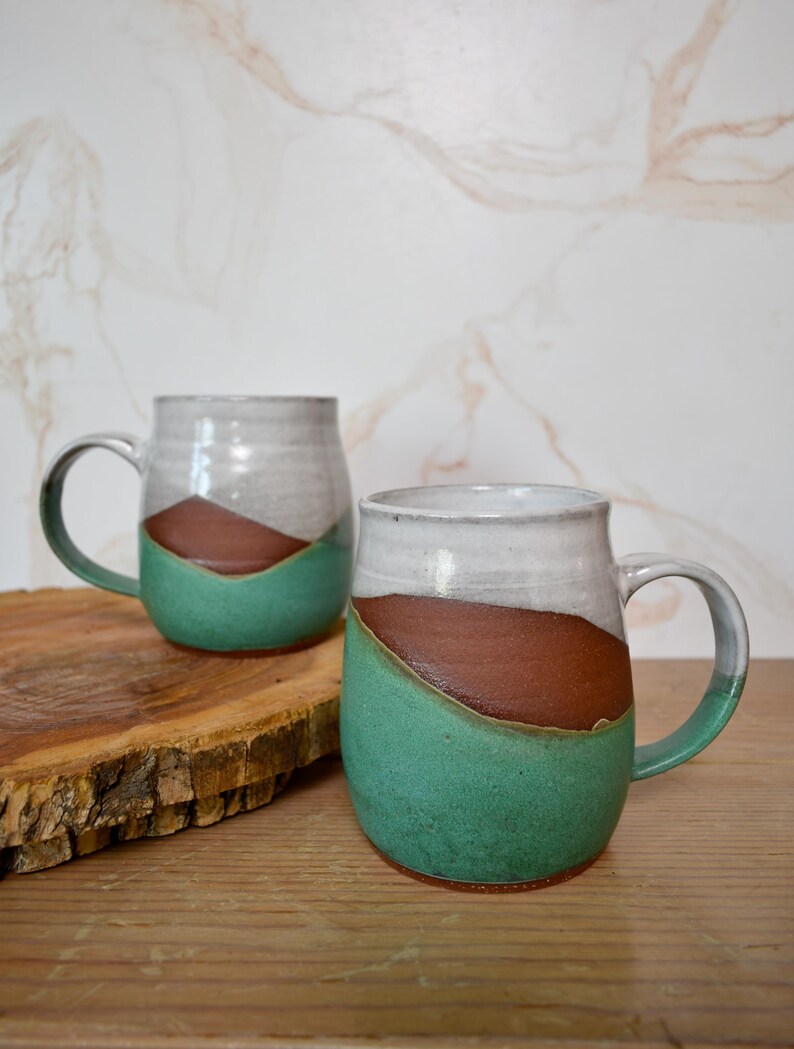 Puede incluir: Dos tazas de cer&aacute;mica hechas a mano con un dise&ntilde;o &uacute;nico. Cada taza presenta una parte superior blanca, una secci&oacute;n media marr&oacute;n y una base verde turquesa. Las tazas tienen asas grandes y se exhiben sobre una superficie de madera.