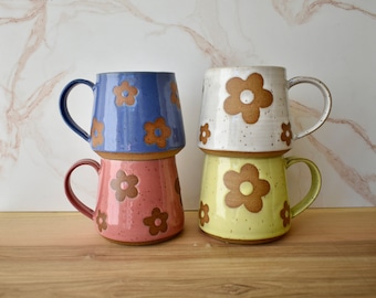 May include: Four ceramic mugs with brown flower designs. The mugs are stacked in two pairs. The top mug is white with brown speckles, the second mug is light blue, the third mug is light yellow, and the bottom mug is pink.