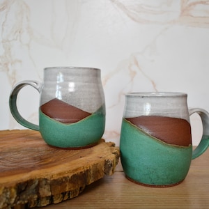 Puede incluir: Dos tazas de cer&aacute;mica hechas a mano con un dise&ntilde;o &uacute;nico. Cada taza presenta una parte superior blanca, una banda diagonal marr&oacute;n y una base verde turquesa. Las tazas est&aacute;n sobre una rodaja de madera, mostrando su naturaleza artesanal.