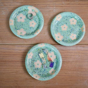 Puede incluir: Tres platos redondos de cerámica para joyas con esmalte verde menta claro, salpicados con puntos azul oscuro y con un patrón repetitivo de flores de color melocotón pálido. Un plato contiene un anillo de plata y otro un par de pendientes colgantes.
