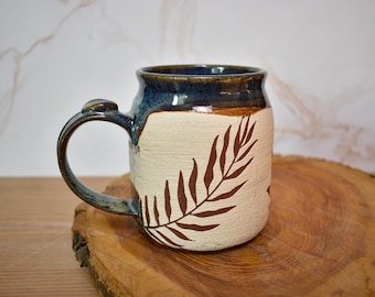 Könnte beinhalten: Eine handgefertigte Keramiktasse mit cremefarbenem Körper und dunkelblauem Rand. Auf der Seite ist ein braunes Farnblattmuster aufgemalt. Die Tasse hat einen großen Henkel und steht auf einer Holzoberfläche.