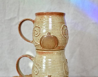 May include: Two stacked ceramic mugs with a speckled cream glaze and a reddish-brown rim. Each mug features a relief design of a central pumpkin surrounded by swirling vine patterns in a matching reddish-brown tone, with a curved handle on the side.