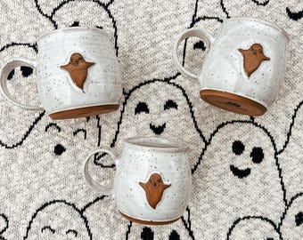 May include: Three white ceramic mugs with speckled surfaces and brown ghost appliqués. Each mug has a handle and a brown base. The mugs are arranged on a gray background with a repeating pattern of smiling ghost faces.