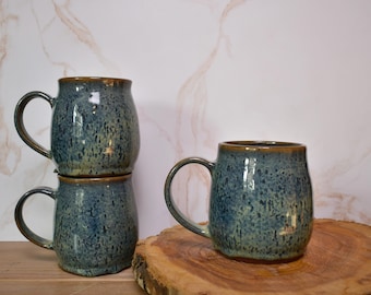 Può includere: Tre tazze in ceramica fatte a mano. Due tazze sono impilate e una è su una superficie di legno. Le tazze sono blu con una glassa macchiata e un bordo e un manico marroni. Le tazze hanno un aspetto rustico e artigianale.