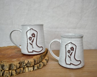 May include: Two white ceramic mugs with a glossy finish, each featuring a brown embossed outline of a cowboy boot decorated with two small flowers. The mugs have a sturdy handle and a slightly tapered shape, showcasing a handcrafted aesthetic.