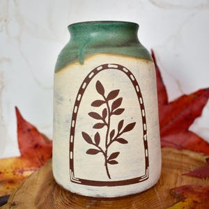 May include: A ceramic vase with a green rim and a cream-colored body. A brown leaf design is centered on the vase. The vase is displayed on a wooden surface with red and yellow leaves.