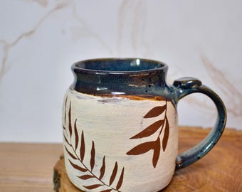 Könnte beinhalten: Eine handgefertigte Keramik-Tasse mit cremefarbenem Korpus und dunkelblauem Rand und Henkel. Die Tasse hat ein braunes Blattmuster. Die Tasse steht auf einer Holzoberfläche.