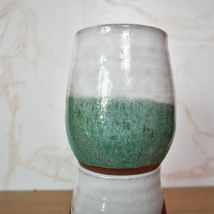 Puede incluir: Un vaso de cer&aacute;mica hecho a mano con un esmalte de dos tonos. La mitad superior es blanca, pasando a un verde texturizado en la parte inferior. El vaso se asienta sobre una base m&aacute;s peque&ntilde;a y similar, mostrando una est&eacute;tica r&uacute;stica y artesanal.