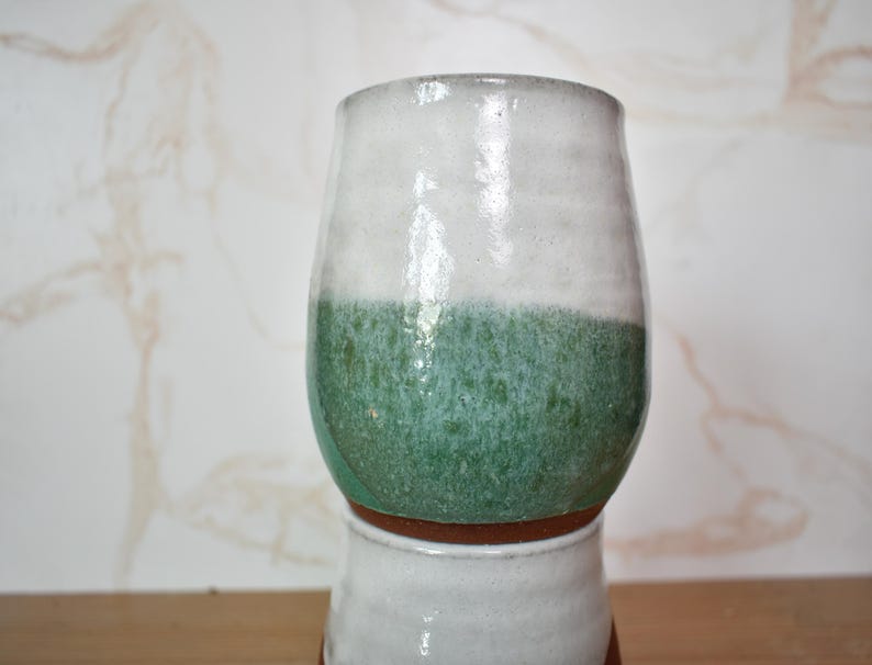 Puede incluir: Un vaso de cer&aacute;mica hecho a mano con un esmalte de dos tonos. La mitad superior es blanca, pasando a un verde texturizado en la parte inferior. El vaso se asienta sobre una base m&aacute;s peque&ntilde;a y similar, mostrando una est&eacute;tica r&uacute;stica y artesanal.