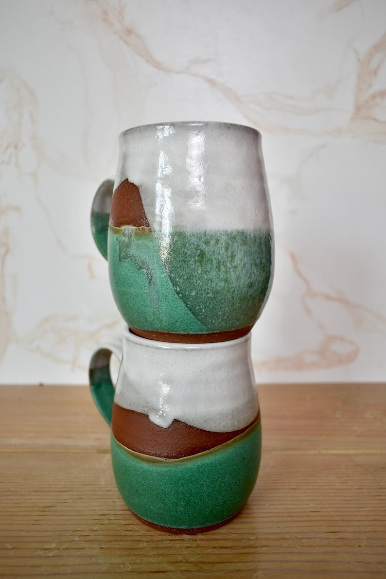 Puede incluir: Dos tazas de cer&aacute;mica hechas a mano apiladas una encima de la otra. Las tazas tienen una parte superior blanca, una secci&oacute;n inferior verde y una banda marr&oacute;n en el medio. Cada taza tiene un asa. Las tazas est&aacute;n sobre una superficie de madera.