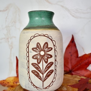 May include: A ceramic vase with a cream-colored body and a green rim. The vase features a brown floral design within a scalloped frame. The vase is displayed on a wooden surface with fall leaves.