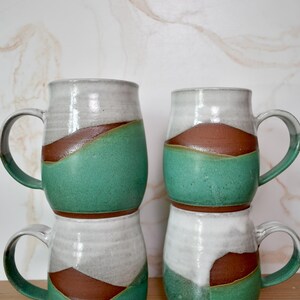 Puede incluir: Cuatro tazas de cer&aacute;mica hechas a mano apiladas. Cada taza presenta un dise&ntilde;o &uacute;nico con una parte superior blanca, una banda marr&oacute;n y una secci&oacute;n inferior verde. Las tazas tienen asas grandes y una est&eacute;tica r&uacute;stica y artesanal.