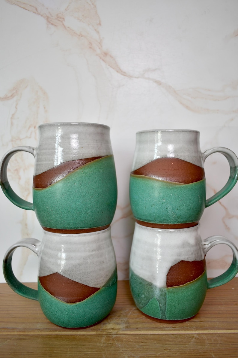 Puede incluir: Cuatro tazas de cer&aacute;mica hechas a mano apiladas. Cada taza presenta un dise&ntilde;o &uacute;nico con una parte superior blanca, una banda marr&oacute;n y una secci&oacute;n inferior verde. Las tazas tienen asas grandes y una est&eacute;tica r&uacute;stica y artesanal.