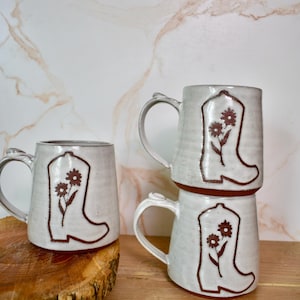 Może przedstawiać: Białe ceramiczne kubki z brązowym wzorem buta kowbojskiego i kwiatów. Jeden kubek stoi na drewnianej powierzchni, a trzy są ułożone w stos. Kubki mają zakrzywiony uchwyt i rustykalny, ręcznie robiony wygląd.