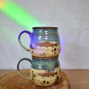 May include: Two stacked ceramic mugs with a rounded shape, featuring a two-tone glaze of blue at the top and a speckled cream at the bottom. Each mug displays a raised, embossed brown fish design on the front and includes a curved handle.