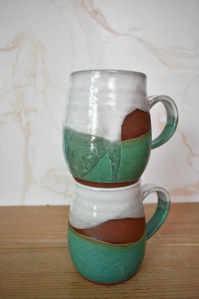 Puede incluir: Dos tazas de cer&aacute;mica hechas a mano apiladas una encima de la otra. Las tazas tienen una parte superior blanca, una secci&oacute;n media marr&oacute;n y una base verde. Cada taza tiene un asa y un acabado brillante. Las tazas est&aacute;n sobre una superficie de madera.