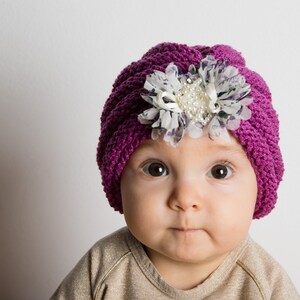 Pode incluir: Um bebê usando um turbante de tricô roxo com um detalhe de flor branca. O turbante tem um enfeite de pérola decorativo.