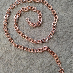 May include: A copper-colored chain necklace with oval links, arranged in a spiral. The necklace has a hook clasp. The chain is displayed on a gray surface.