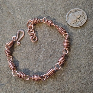 May include: A handmade copper bracelet with a spiral design. The bracelet features a series of coiled copper beads connected by small links. It has a hook clasp for closure. A US nickel is next to the bracelet for size comparison.