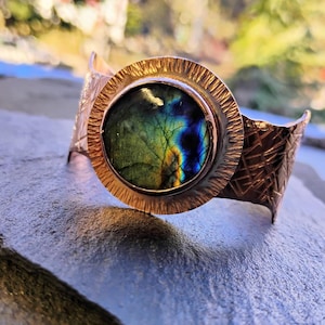 May include: A copper-colored cuff bracelet with a large, round, iridescent gemstone at its center. The gemstone displays a mix of green, blue, and yellow hues. The bracelet has a textured design around the gemstone and on the cuff.