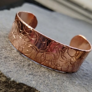 May include: A copper cuff bracelet with a repeating circular pattern. The bracelet has a wide band and open ends. The metal has a warm, reddish-brown hue and a polished finish. The design is etched into the surface.