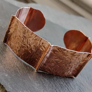 May include: A copper cuff bracelet with a textured surface. The bracelet has a rectangular shape with rounded edges and a warm, reddish-brown color. The cuff is open at the top and bottom.