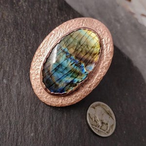 May include: Oval-shaped copper pendant with a large, iridescent labradorite stone. The stone displays blue, green, and yellow hues. The copper setting has a textured, hammered appearance. A US nickel coin is next to the pendant for size comparison.
