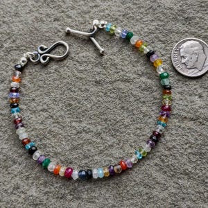 May include: A multi-colored beaded bracelet featuring small, faceted, translucent gemstones in various hues including red, blue, green, yellow, and purple. The bracelet has a silver-toned toggle clasp and is shown next to a US dime for scale.
