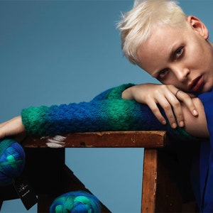 May include: A woman with short, platinum blonde hair is wearing a blue and green sweater and a blue dress. She is leaning on a wooden step stool with a ball of blue and green yarn in her hand.