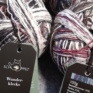 May include: Close-up of several skeins of yarn in shades of gray, white, and burgundy. The yarn is wound into neat balls, with tags attached. The tags feature the brand logo and the text "Wunderklecks".
