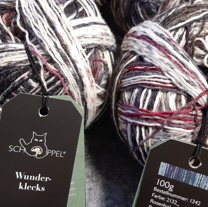 May include: Close-up of several skeins of yarn in shades of gray, white, and burgundy. The yarn is wound into neat balls, with tags attached. The tags feature the brand logo and the text "Wunderklecks".