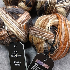 May include: Four balls of yarn in shades of brown, beige, and white. The yarn is arranged in a circle, with the balls overlapping. The yarn is labeled "Schöppel Wunder-klecks 100g Bestellnummer: 1342 Farbe: 2151_ Vollmilch-T Partin"