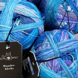 May include: Three balls of blue, purple, and white yarn with a black tag that reads "Schoppel Wunder-klecks 100g Bestelinummer: 1342 Farbe: 2431 Fuji lights Partie: 729809"