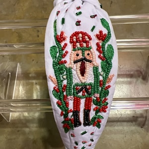 Puede incluir: Diadema de tela blanca con un diseño de cascanueces con cuentas. El cascanueces tiene un sombrero rojo y blanco, un bigote negro y detalles en rojo y verde. La diadema está decorada con cuentas rojas y verdes.