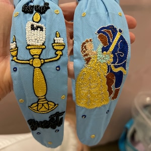 Op de afbeelding: Twee blauwe stoffen hoofdbanden met geborduurde ontwerpen. Een hoofdband heeft een gele en gouden kandelaar met een witte parel-vlam en het woord "Sweet" in het zwart. De andere hoofdband heeft een scène van Belle en het Beest uit Belle en het Beest.