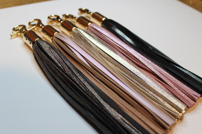 Pu&ograve; includere: Un set di sei portachiavi con anelli in metallo dorato e nappe in pelle di diversi colori. Le nappe sono nere, argentate, beige, rosa chiaro, dorate e rosa.