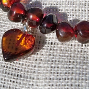 May include: A heart-shaped amber pendant hangs from a strand of amber beads. The beads are a deep reddish-brown color and have a glossy finish. The pendant is a lighter shade of amber and has a more translucent appearance.