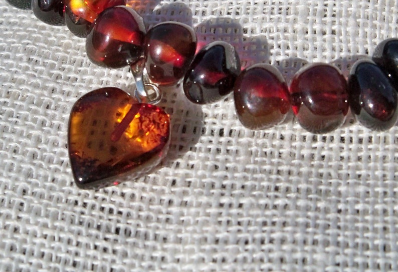 May include: A heart-shaped amber pendant hangs from a strand of amber beads. The beads are a deep reddish-brown color and have a glossy finish. The pendant is a lighter shade of amber and has a more translucent appearance.