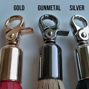 Pu&ograve; includere: Tre portachiavi con chiusure metalliche e nappine di diversi colori. Il portachiavi a sinistra ha una chiusura dorata e una nappina rossa. Il portachiavi al centro ha una chiusura color metallo canna di fucile e una nappina beige. Il portachiavi a destra ha una chiusura argentata e una nappina nera.