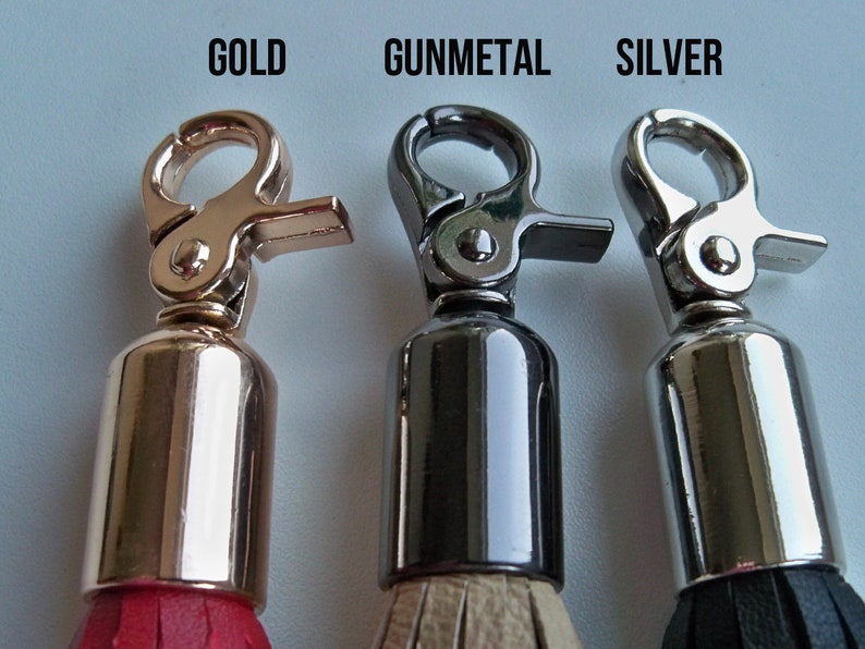 Pu&ograve; includere: Tre portachiavi con chiusure metalliche e nappine di diversi colori. Il portachiavi a sinistra ha una chiusura dorata e una nappina rossa. Il portachiavi al centro ha una chiusura color metallo canna di fucile e una nappina beige. Il portachiavi a destra ha una chiusura argentata e una nappina nera.