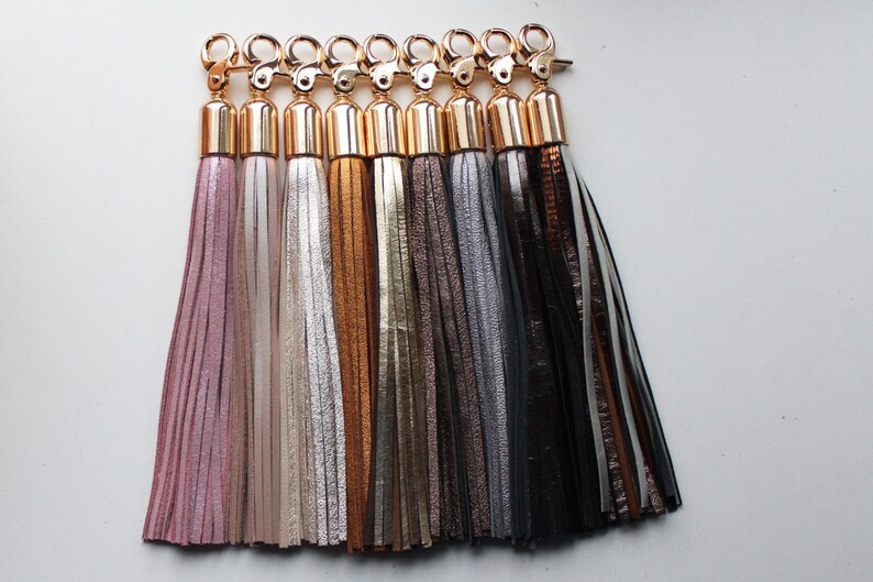 Pu&ograve; includere: Un set di otto portachiavi con nappe in vari colori, tra cui rosa, beige, oro, marrone, grigio e nero. Ogni portachiavi ha un anello metallico dorato e un cappuccio metallico dorato.