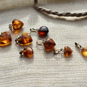 May include: A collection of eight amber heart-shaped pendants with silver bail settings. The pendants are arranged on a white fabric background.