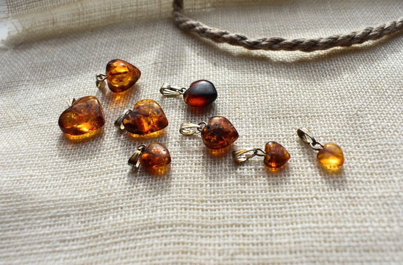 May include: A collection of eight amber heart-shaped pendants with silver bail settings. The pendants are arranged on a white fabric background.