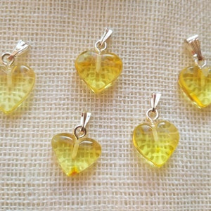 May include: Five yellow heart-shaped amber pendants with silver bail loops. The pendants are arranged in a semi-circle on a beige fabric background.