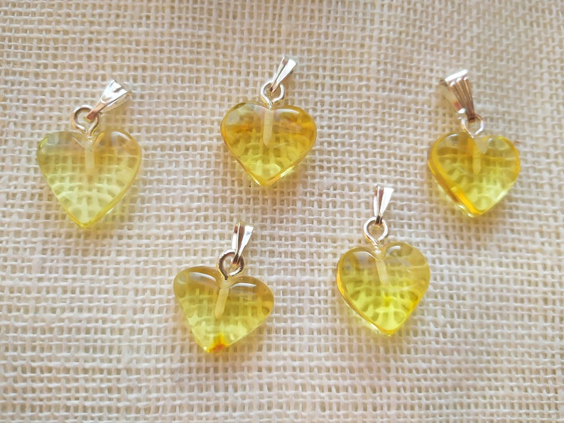 May include: Five yellow heart-shaped amber pendants with silver bail loops. The pendants are arranged in a semi-circle on a beige fabric background.