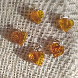 May include: Five amber heart-shaped pendants with silver bail loops. The pendants are arranged in a circle on a beige fabric background.